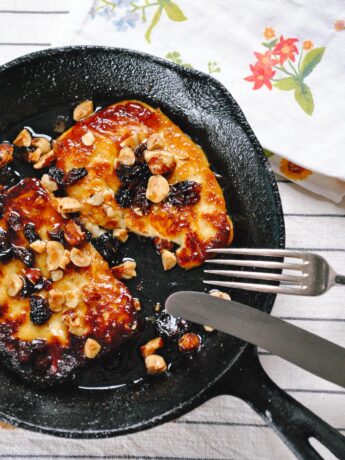 A small cast iron pan with two thick glossy slices of haloumi in it garnished with currants and hazelnuts