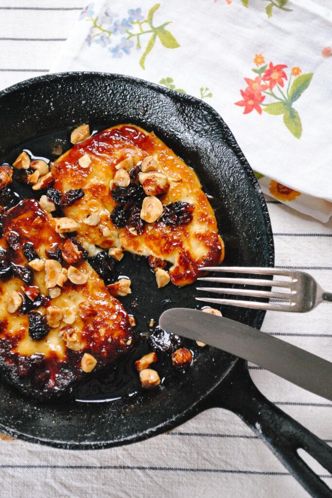 A small cast iron pan with two thick glossy slices of haloumi in it garnished with currants and hazelnuts