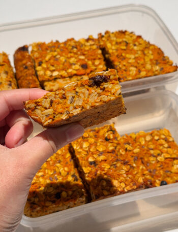 Hand holding a slice of orange coloured oat slice flecked with raisins.