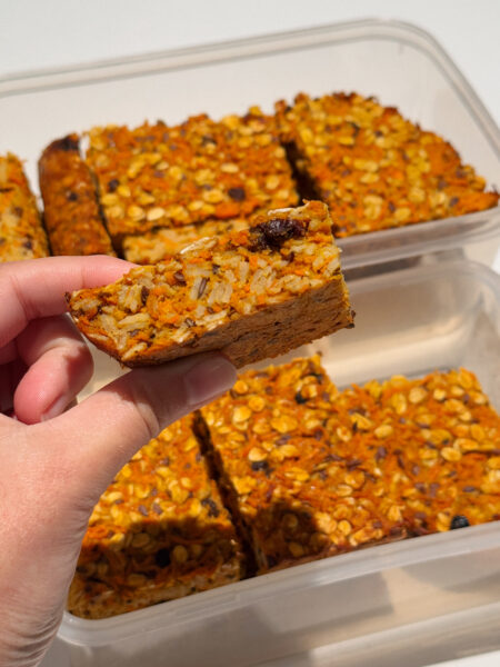 Hand holding a slice of orange coloured oat slice flecked with raisins.