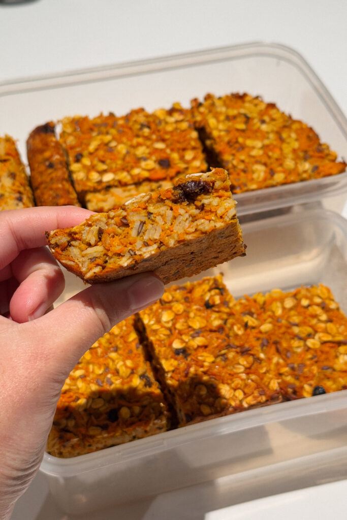 Hand holding a slice of orange coloured oat slice flecked with raisins.